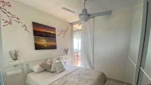 Une chambre à coucher avec un lit, un ventilateur de plafond, une fenêtre avec des rideaux et un tableau accroché au mur.