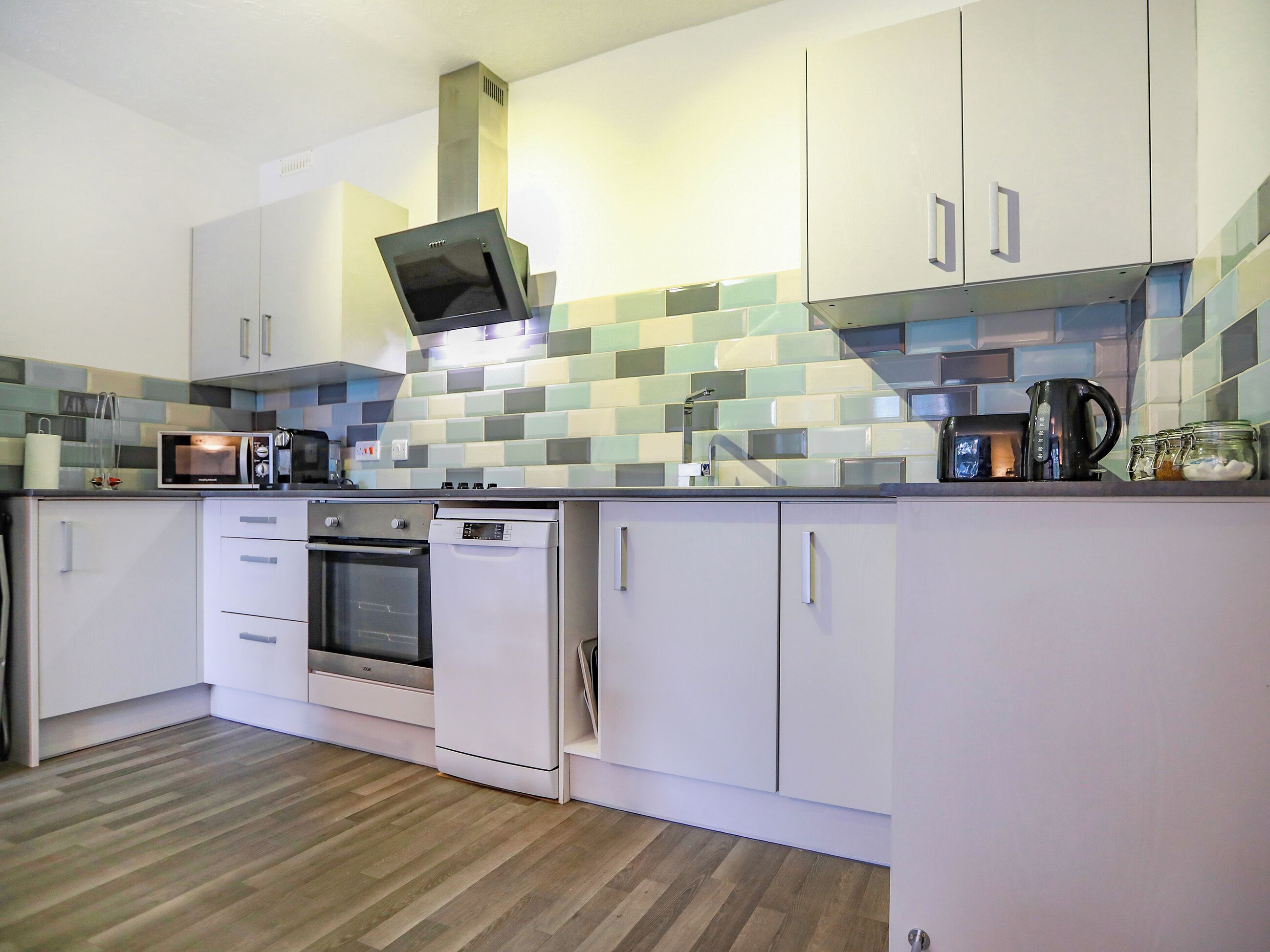 A modern kitchen with white cabinets, a stainless steel range hood, a built-in oven, a dishwasher, and a tiled backsplash.
