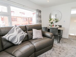 A modern living room with a leather sofa, a dining table with chairs, and a large window with a view of a red brick building. - Daisy's Den (Whitby)