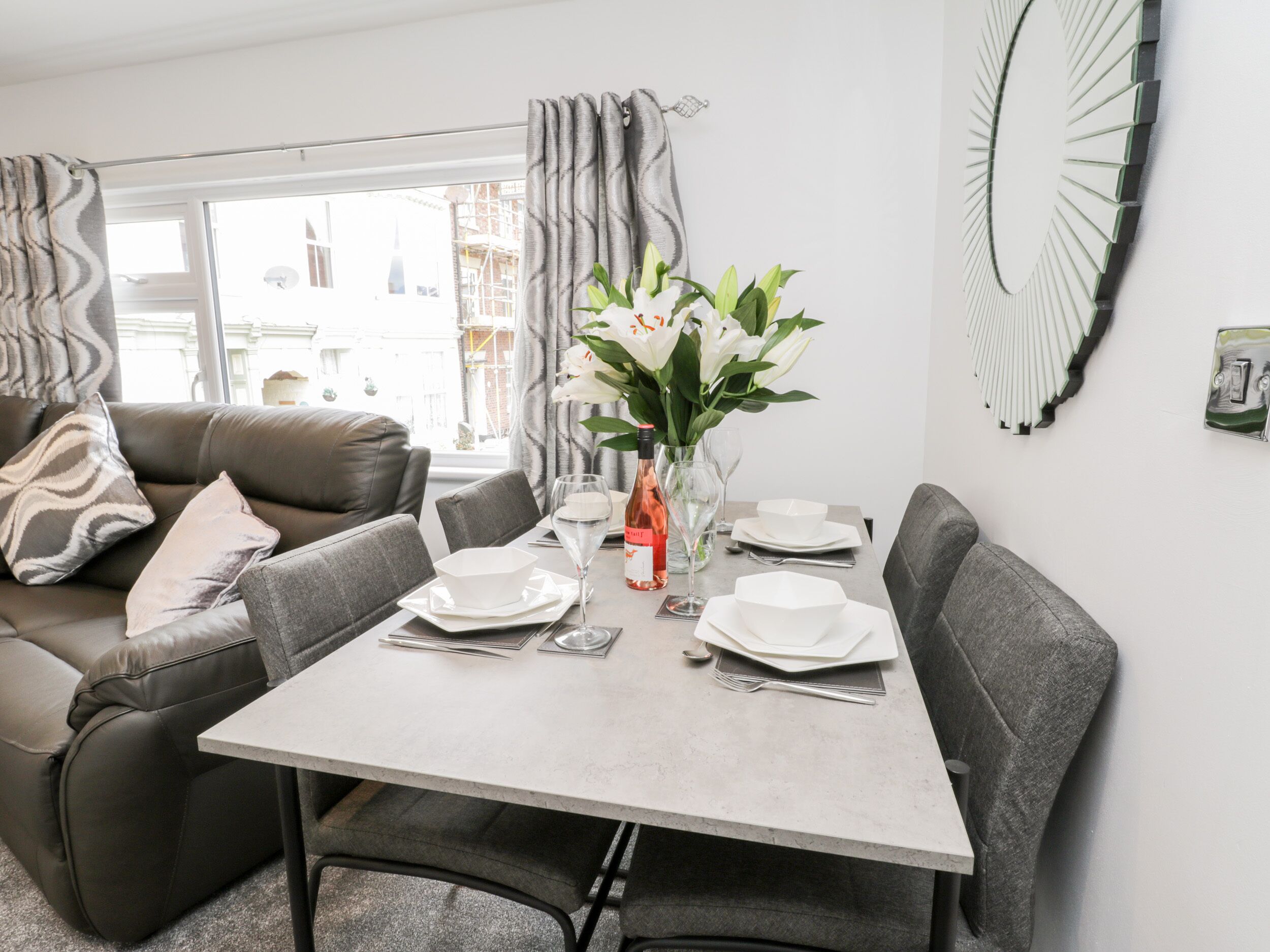 A dining area with a table set for four, a vase with flowers, and a view of the city through large windows.