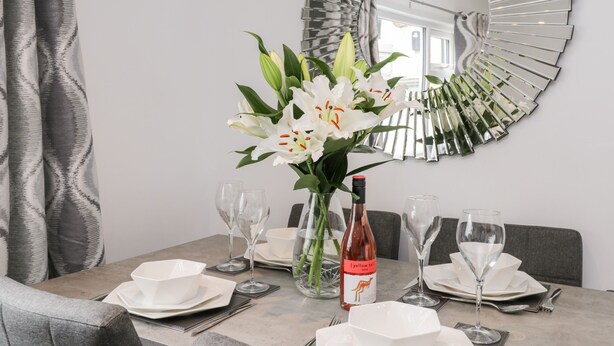 Une table à manger dressée avec de l’assiette blanche, des bols et des verres à vin, une bouteille de vin et un vase rempli de fleurs blanches.