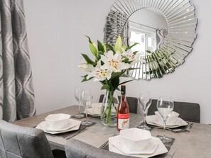 A dining table set with white plates, bowls, and wine glasses, a bottle of wine, and a vase with white flowers. - Daisy's Den (Whitby)
