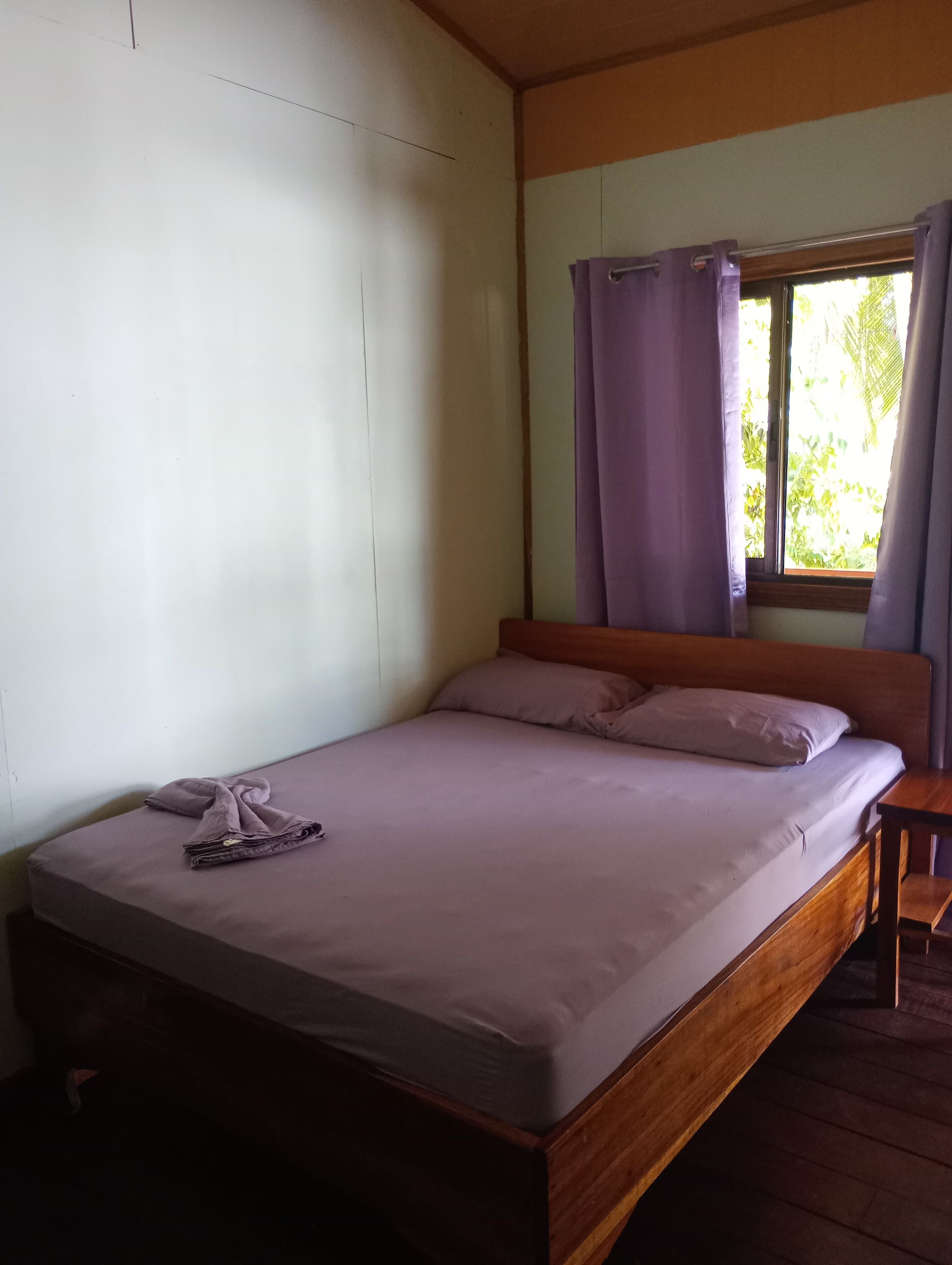 Ein Einzelbett mit Holzrahmen, ein kleiner Nachttisch und ein Fenster mit violetten Vorhängen.