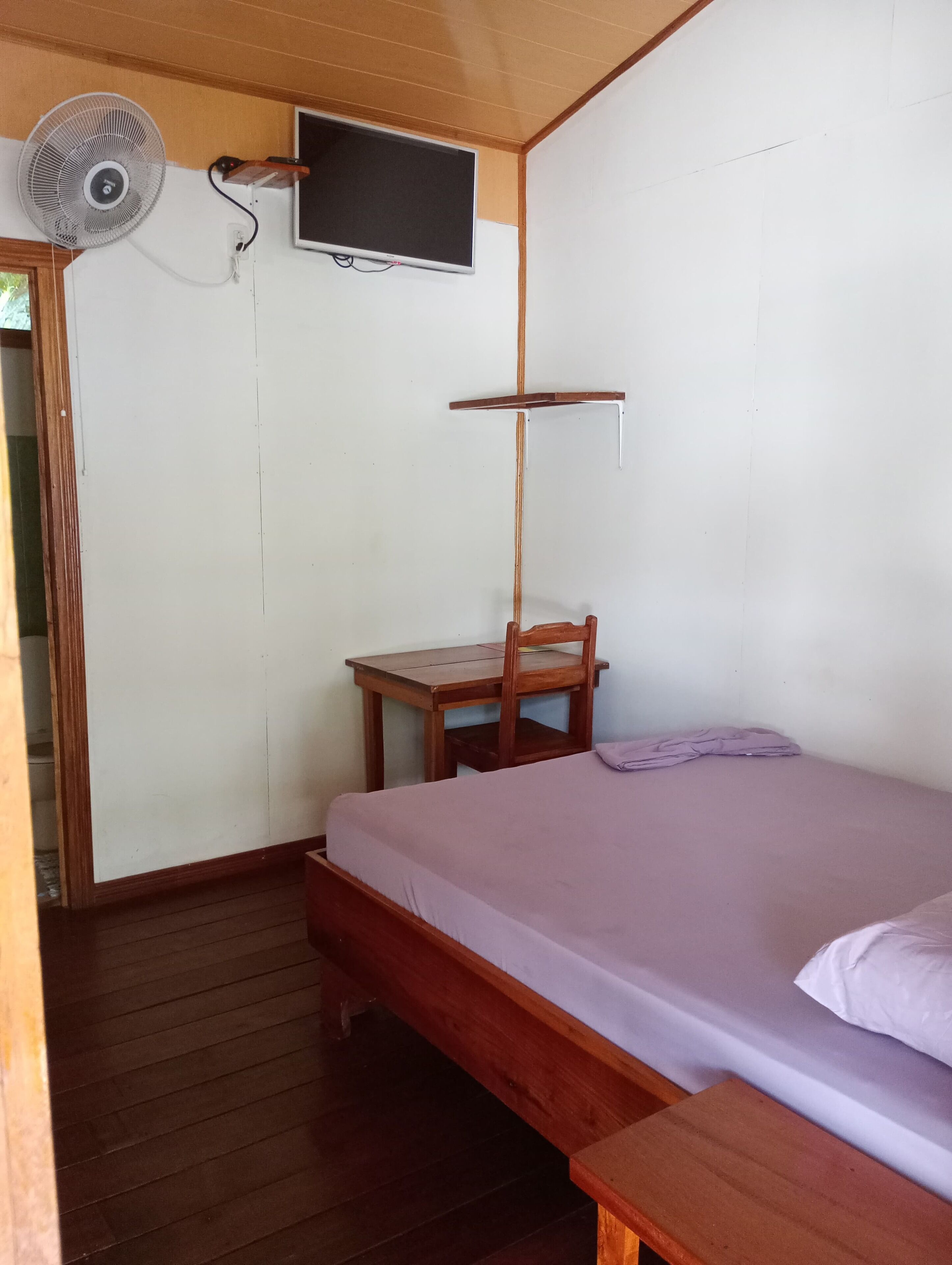 A simple room with a bed, a small wooden table, a chair, a wall-mounted TV, and a ceiling fan.