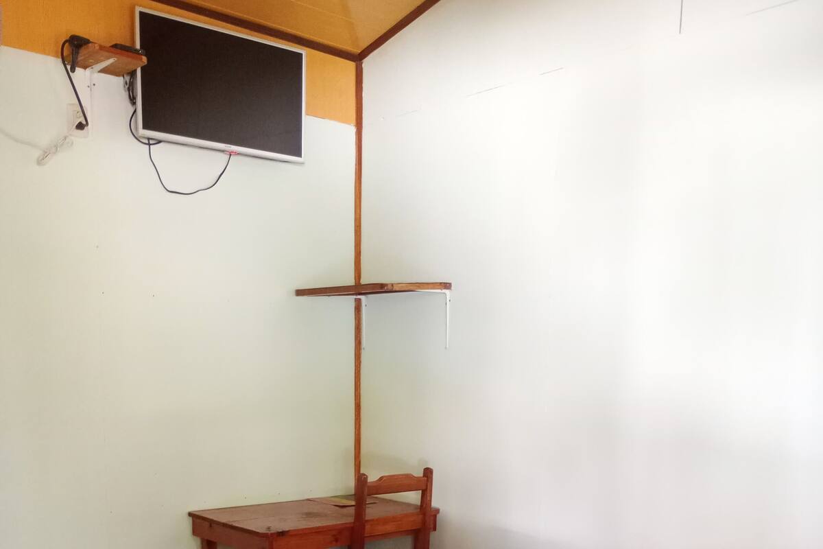 Una habitación pequeña y sencilla con una cama de madera, un escritorio con una silla y un televisor fijado a la pared.