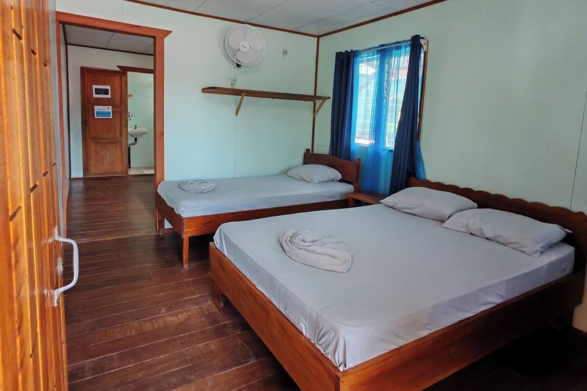 Una habitación con dos camas de madera, un ventilador de techo y una puerta que da a un baño.