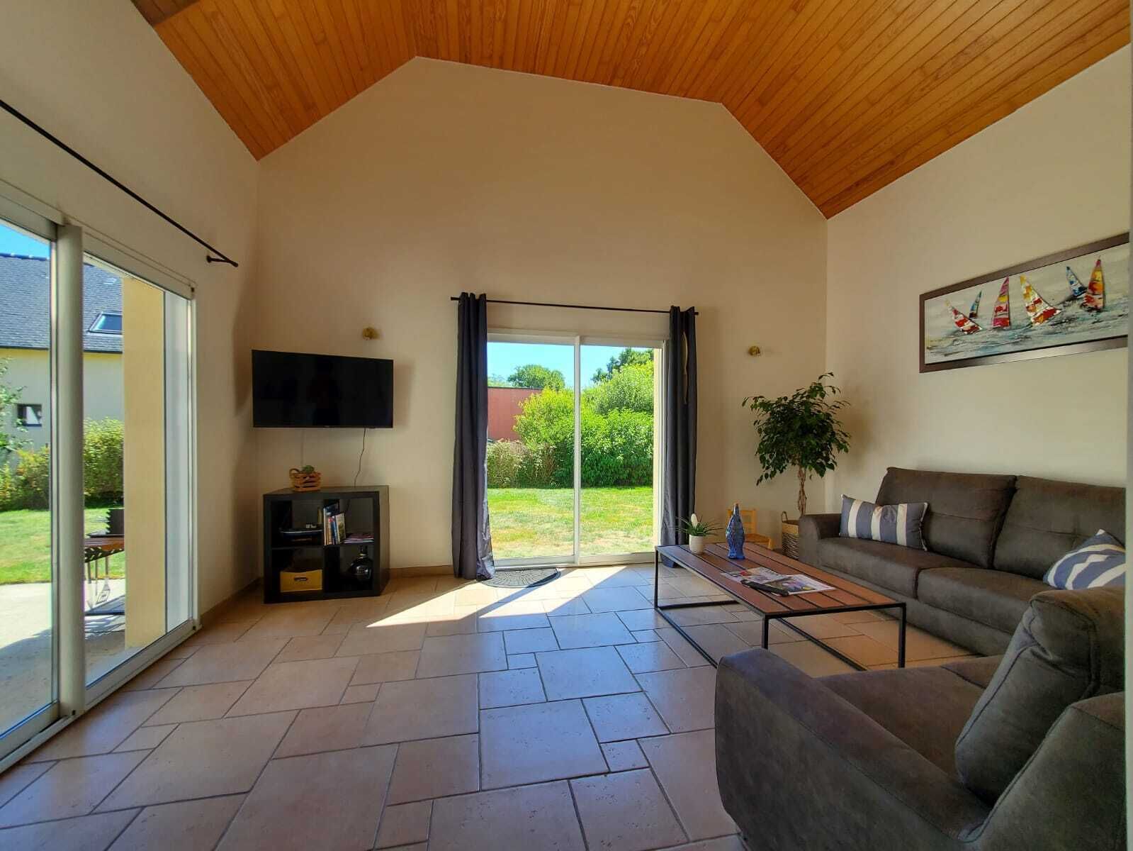 Uma sala de estar espaçosa com uma grande porta de vidro deslizante que dá para um jardim, uma TV de tela plana na parede, um sofá marrom e uma mesa de centro.