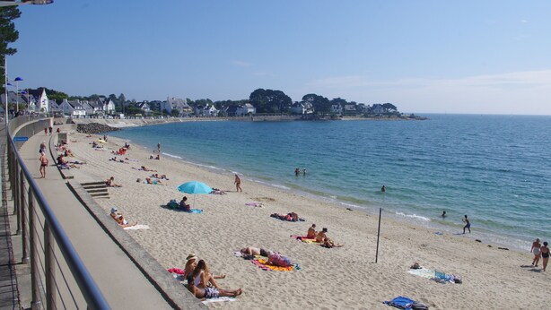 Ein Strand mit sonnenbadenden und schwimmenden Personen, eine Promenade mit Sonnenschirmen und Häuser im Hintergrund.