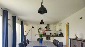 Eine moderne Küche mit weißen Schränken, ein Essbereich mit einem Holztisch und dunklen Stühlen sowie ein großes Fenster mit blauen Vorhängen.