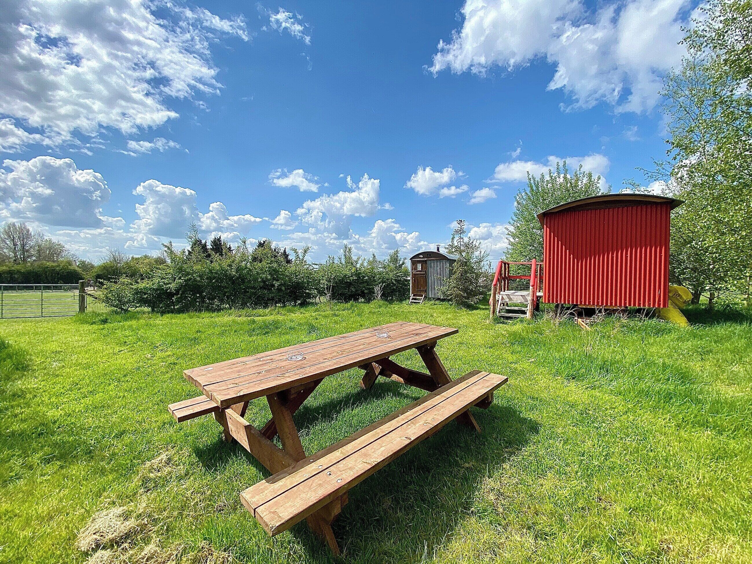 Una mesa de picnic en un área verde con un cobertizo rojo y una pequeña construcción de madera al fondo.