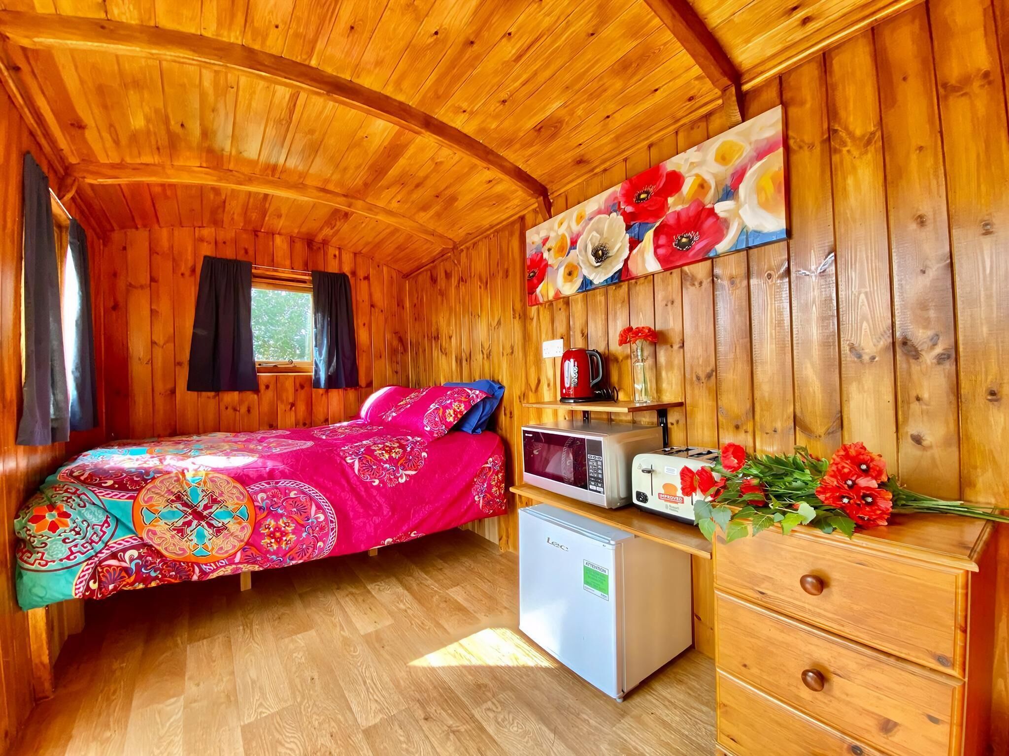 A wooden cabin interior with a bed, a small refrigerator, a microwave, a toaster, and a wall-mounted shelf with a kettle and flowers.