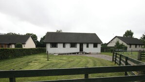 Um cenário rural com uma casa branca e teto escuro, cercada por uma área gramada e uma cerca de madeira.