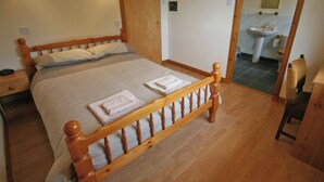 Uma cama bem arrumada com almofadas e toalhas dobradas, uma mesa de cabeceira de madeira, uma porta de madeira e vista para um banheiro com pia e espelho.