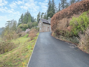 A stone building with a paved driveway leading to it, surrounded by trees and shrubs.