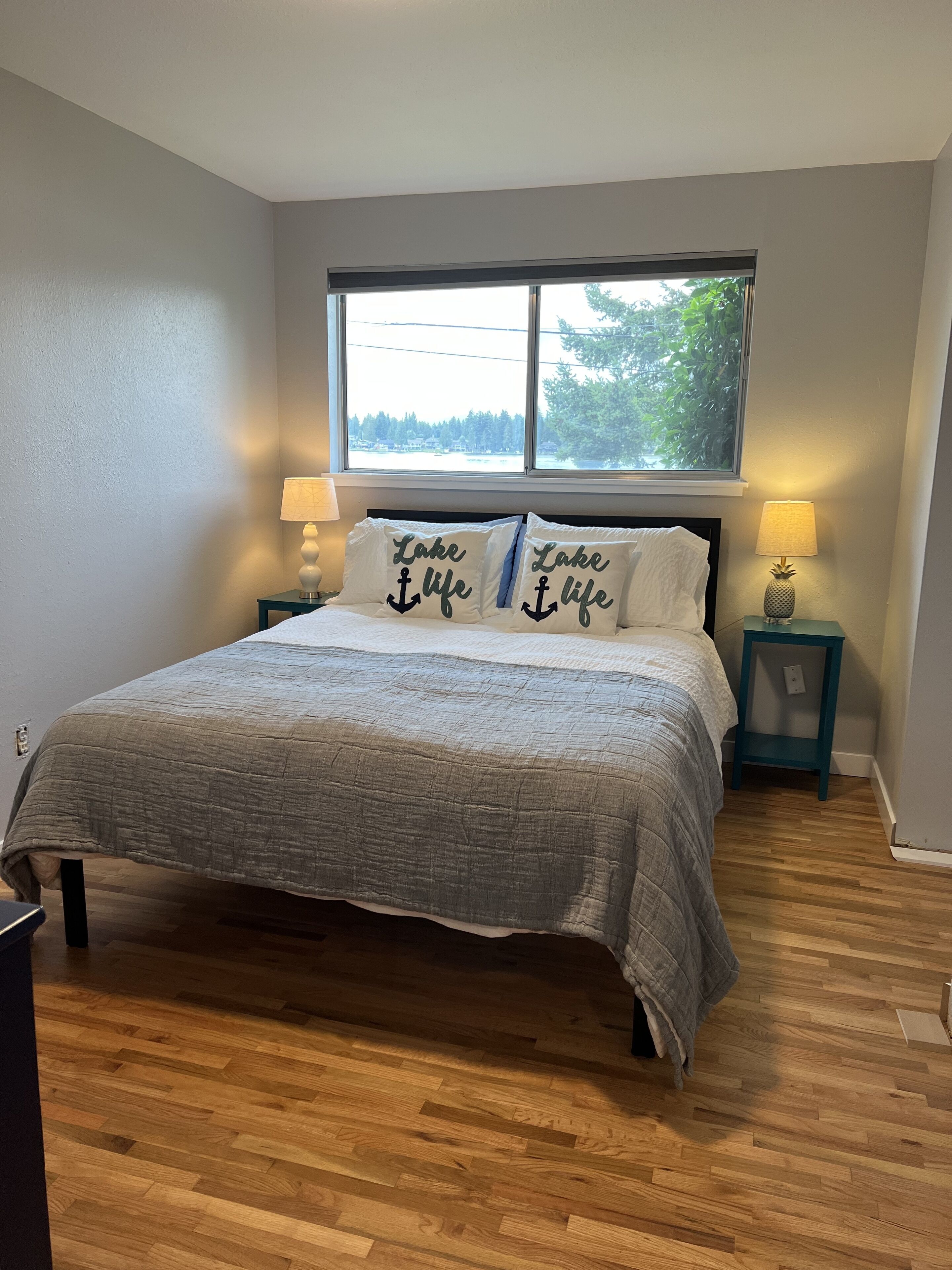 A bedroom with a bed, two nightstands, a window with a view of trees, and two pillows with the text "Lake life".