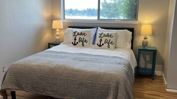 A bedroom with a bed, two nightstands, a window with a view of trees, and two pillows with the text "Lake life".