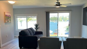 Ein geräumiges Wohnzimmer mit einem dunkelgrauen Sofa, zwei Sesseln und einem Deckenventilator. Große Fenster bieten Blick auf ein Gewässer und viel Grün.