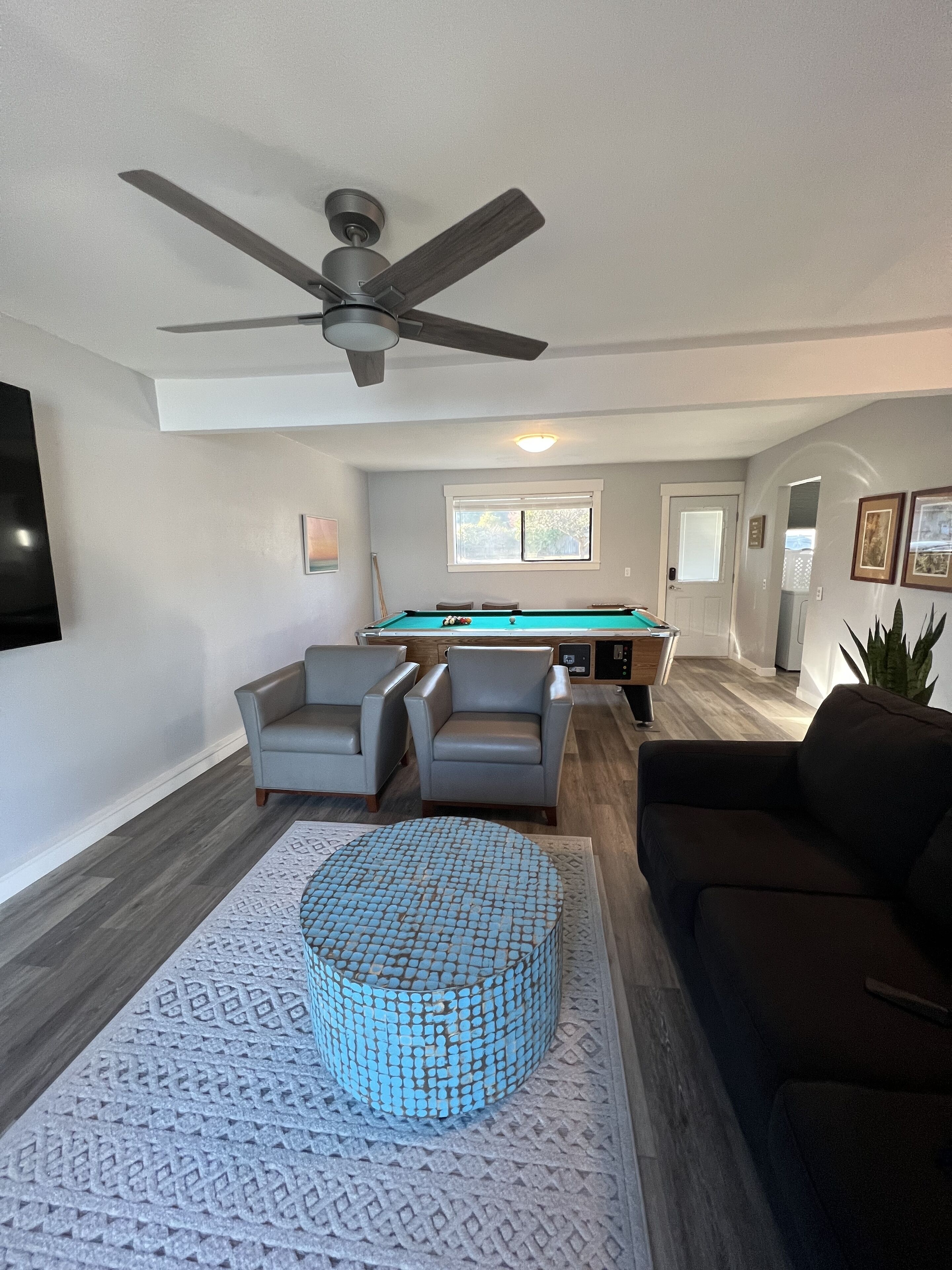 A spacious living room with a pool table, two armchairs, a dark sofa, and a ceiling fan.