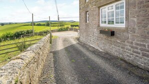 Un bâtiment en pierre avec une allée de gravier, une clôture en bois et un paysage rural en arrière-plan.