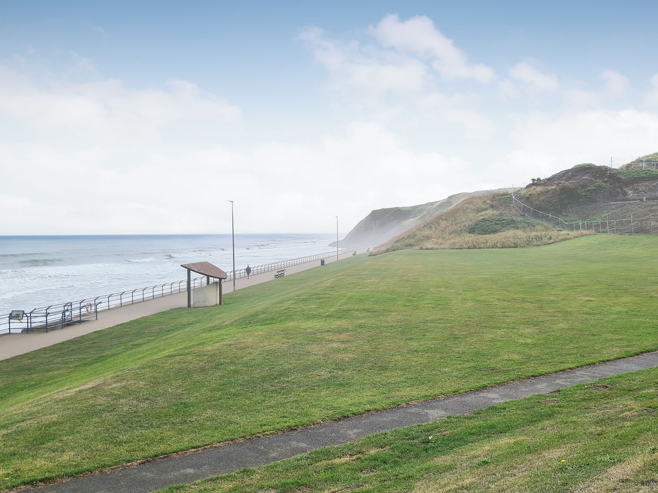 Un parc situé en bord de mer, doté d’une pelouse, d’une promenade dallée et d’un banc.