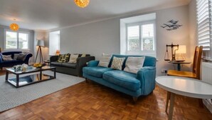 A spacious living room with a blue sofa, a coffee table, and a lamp. There are windows with blinds and a decorative wall piece. - Amroth Holiday home with Sea Views (Narberth)