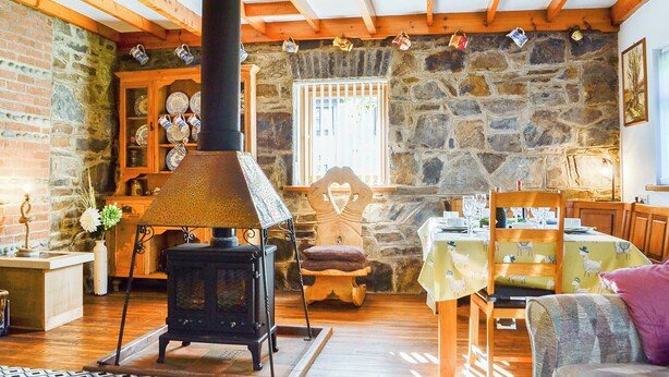 Une pièce chaleureuse avec un mur en pierre, des poutres apparentes, un poêle noir, une table en bois et une chaise.