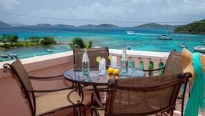 A round glass table with wicker chairs, a glass bottle, and a glass of water on a balcony overlooking a turquoise sea with boats. - GRANDE BAY RESORT - 3BR/ 2BA Directly on Cruz Bay (St John)