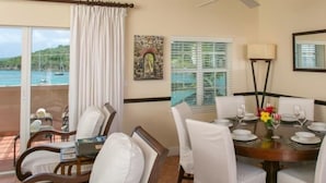 A dining area with a round table set for four, white cushioned chairs, a vase with flowers, and a view of the sea through a window. - GRANDE BAY RESORT - 3BR/ 2BA Directly on Cruz Bay (St John)