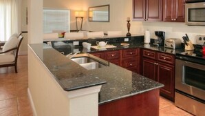 A kitchen with dark wooden cabinets, a granite countertop, stainless steel appliances, and a dining area with a chair and table. - GRANDE BAY RESORT - 3BR/ 2BA Directly on Cruz Bay (St John)