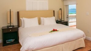 A neatly made bed with white linens and a beige bed skirt, a bedside table with a lamp, and a view of the sea through a balcony door. - GRANDE BAY RESORT - 3BR/ 2BA Directly on Cruz Bay (St John)