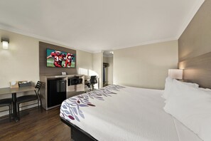 A hotel room with a bed, desk, chair, refrigerator, microwave, and a TV mounted on the wall.