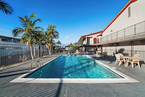 Ein rechteckiger Swimmingpool mit klarem, blauem Wasser, umgeben von einer gefliesten Liegefläche. Auf der Liegefläche stehen Liegestühle und ein Tisch mit Stühlen.