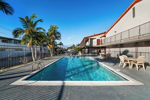 Una piscina rectangular con agua cristalina y azul, rodeada por una terraza con baldosas. Hay sillones de descanso y una mesa con sillas en la terraza.