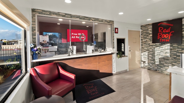 A reception area with a Red Roof Inn sign, a reception desk, a red chair, and a view of the street outside. - Red Roof Inn Los Angeles – Bellflower (Bellflower)