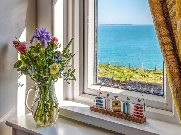 A clear view of the sea through a window, a vase with flowers on a windowsill, and a small wooden dock model.