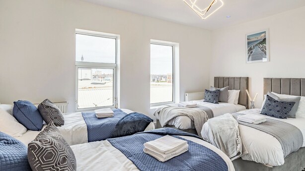 Ein Hotelzimmer mit drei Betten, großen Fenstern, einer modernen Leuchte und Blick auf die Stadt.