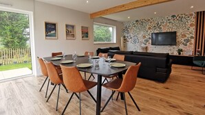 A modern living room with a dining area, a black sofa, a flat-screen TV, and a large window overlooking a garden.