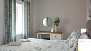 A bedroom with a bed, a bedside table, a nightstand, a dresser, a mirror, and a window with curtains. - Sunrise Cottage - UK35813 (Llanddulas, near Llandudno)