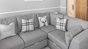 A grey sectional sofa with plaid cushions, a framed artwork of birds, and a wooden door in a neutral-toned room. - Sunrise Cottage - UK35813 (Llanddulas, near Llandudno)