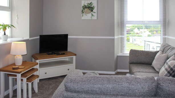 A modern living room with a grey sofa, a television on a wooden stand, a small table with a lamp, and a window with blinds. - Sunrise Cottage - UK35813 (Llanddulas, near Llandudno)