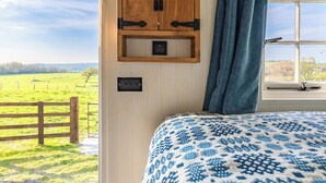 Una camera da letto con una grande finestra, uno scaffale in legno con libri, una coperta a motivi decorativi e vista su un campo verde.