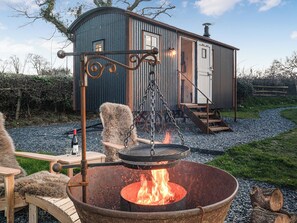 A rustic outdoor setting with a metal fire pit, a wooden bench, and a small metal building with a door and steps.