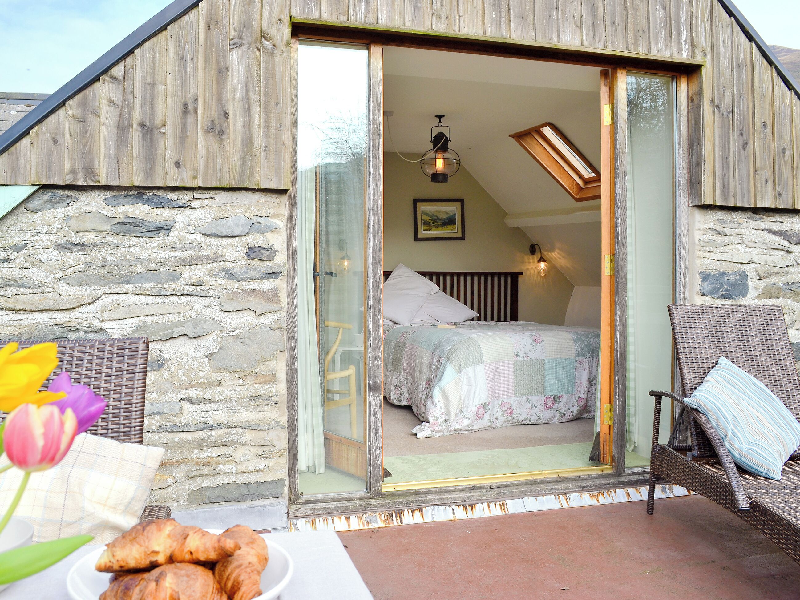 A rustic bedroom with a bed, a wicker chair, and a breakfast setup with croissants.