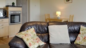 A living room with a leather sofa, floral cushions, a small table with a lamp, and a kitchen area with appliances.