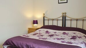 A neatly made bed with a floral bedspread and a purple blanket, a bedside table with a lamp, and a framed picture on the wall.