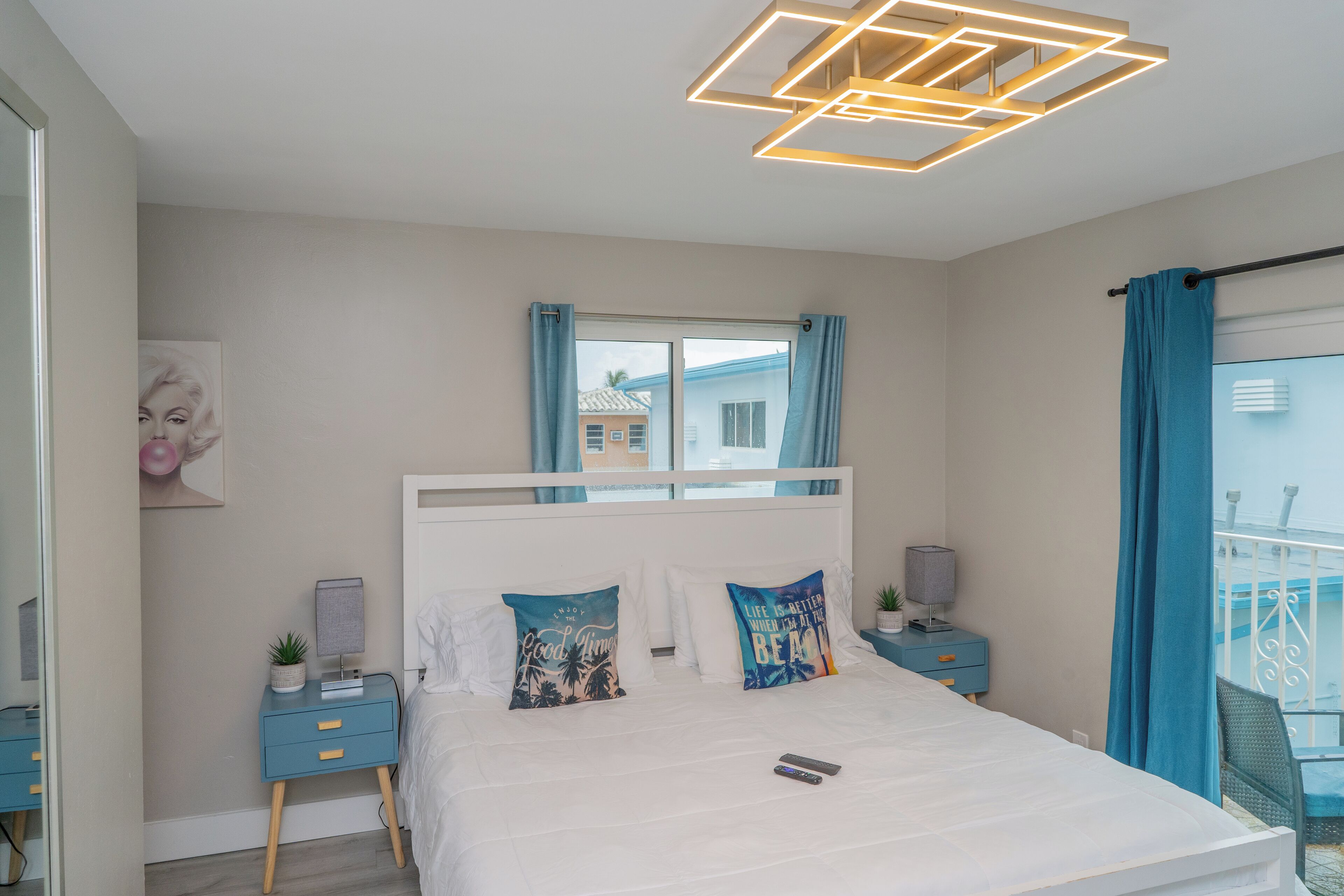 Une chambre moderne avec un lit blanc, deux coussins décoratifs, une table de chevet avec une lampe, une petite plante et une fenêtre avec des rideaux.