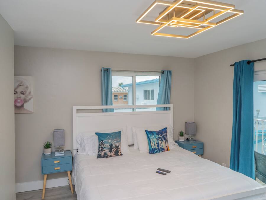 Une chambre moderne avec un lit blanc, deux coussins décoratifs, une table de chevet avec une lampe, une petite plante et une fenêtre avec des rideaux.