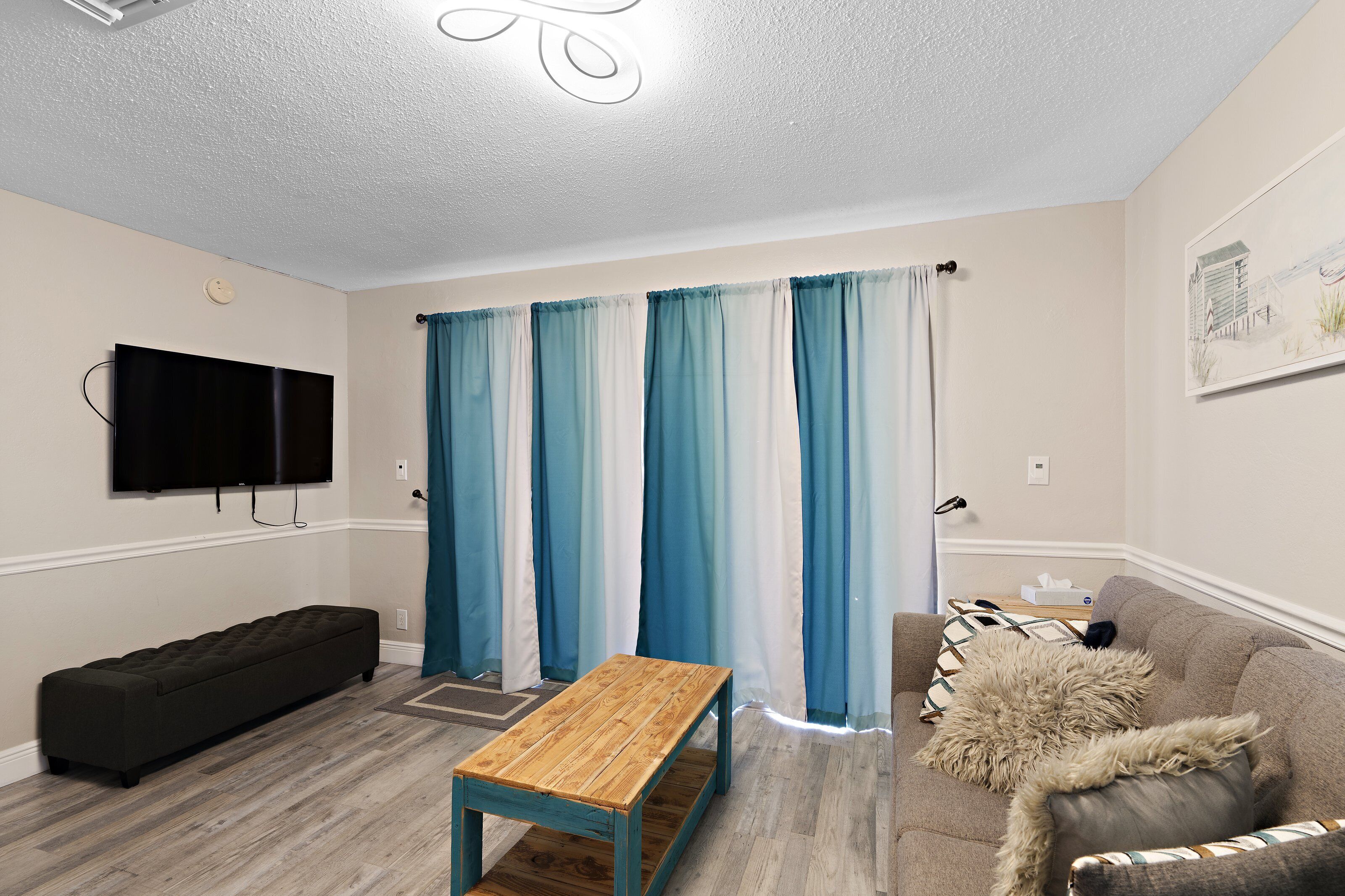 A living room with a flat-screen TV mounted on the wall, a wooden bench, a sofa with a furry throw, and a window with blue and white curtains.
