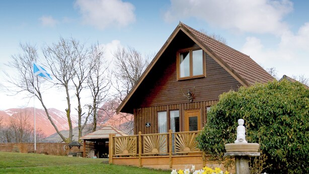 Une maison en bois avec un balcon, entourée d’un jardin où poussent des jonquilles, et un drapeau en arrière-plan.