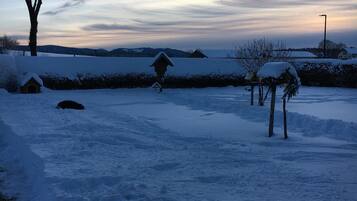 A snowy landscape with a wooden structure, a tree, and a distant view of hills.